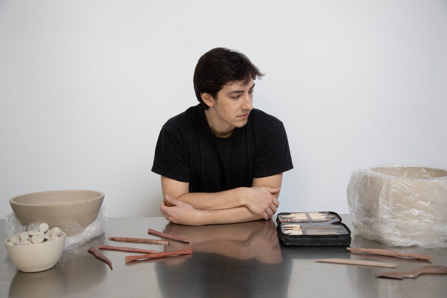 Dapo seated at a wide studio table surrounded by unfinished clay pieces and tools