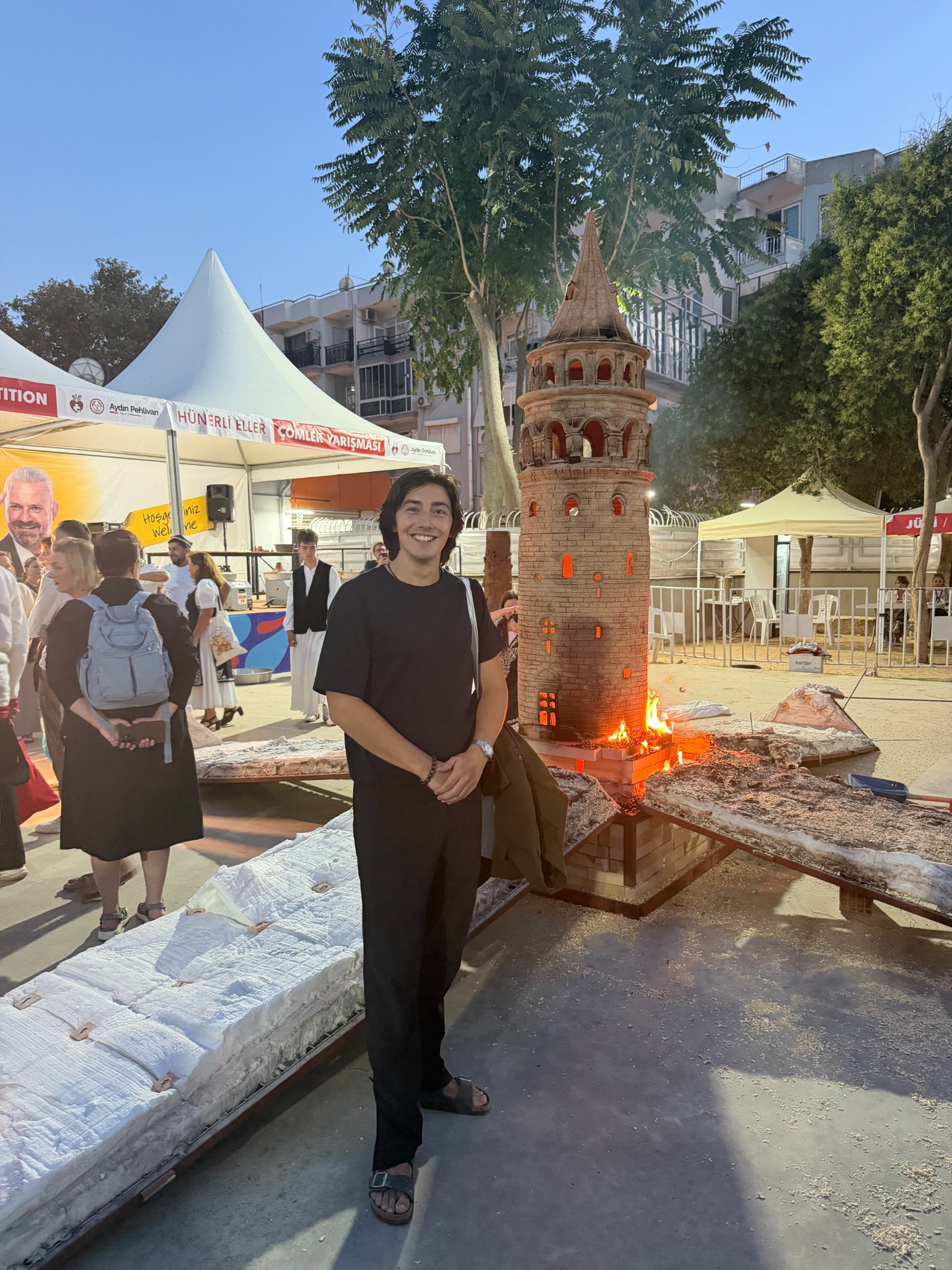 A detailed ceramic tower being fired at night with Daniel standing beside it